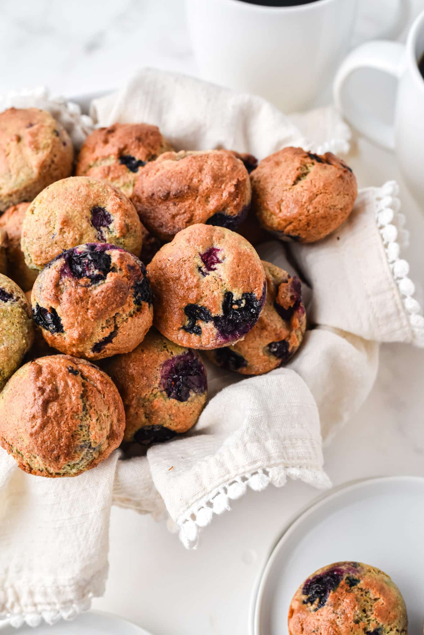 Mini muffins de arándanos en una canasta forrada con una toalla junto a una taza de café.