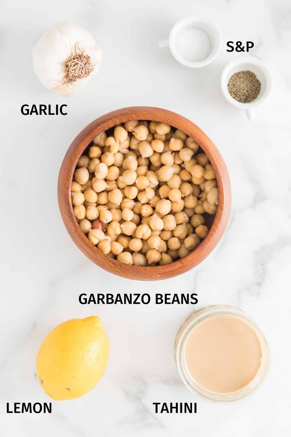 Ingredientes para hacer hummus sin aceite en cuencos pequeños sobre una superficie blanca.