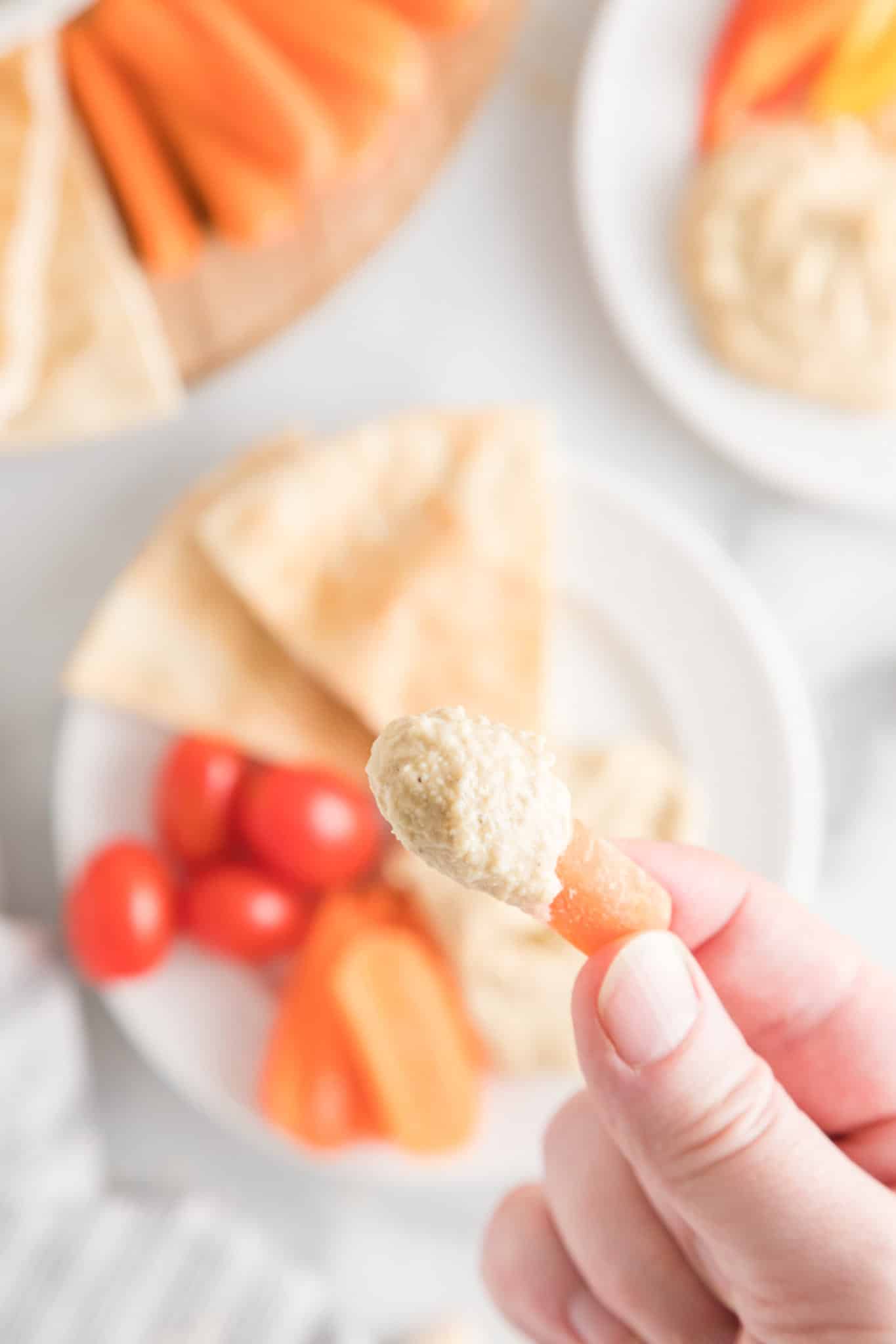 Primer plano de una mano que sostiene una zanahoria pequeña que ha sido sumergida en hummus casero.