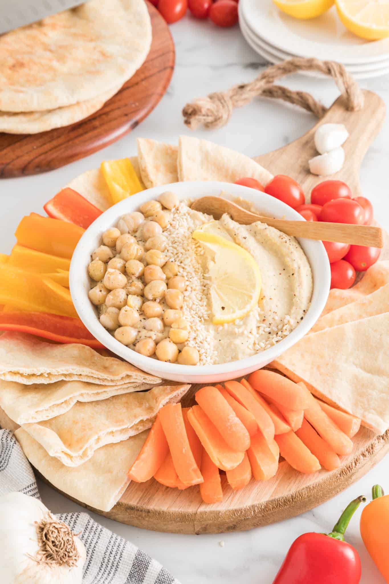 Una fuente de madera con verduras en rodajas y pita en cuartos alrededor de un tazón pequeño de hummus.