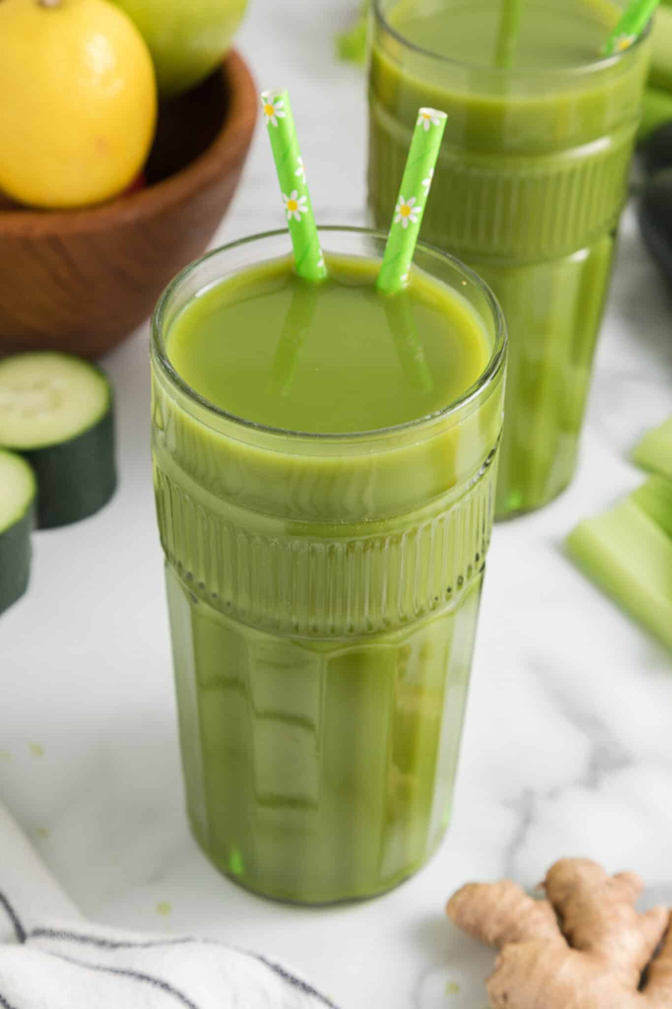 Un vaso alto de jugo verde de limón y jengibre con dos pajitas.